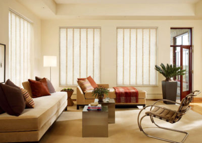 cream colored light filtering large vane vertical blinds in living room with red and brown accents
